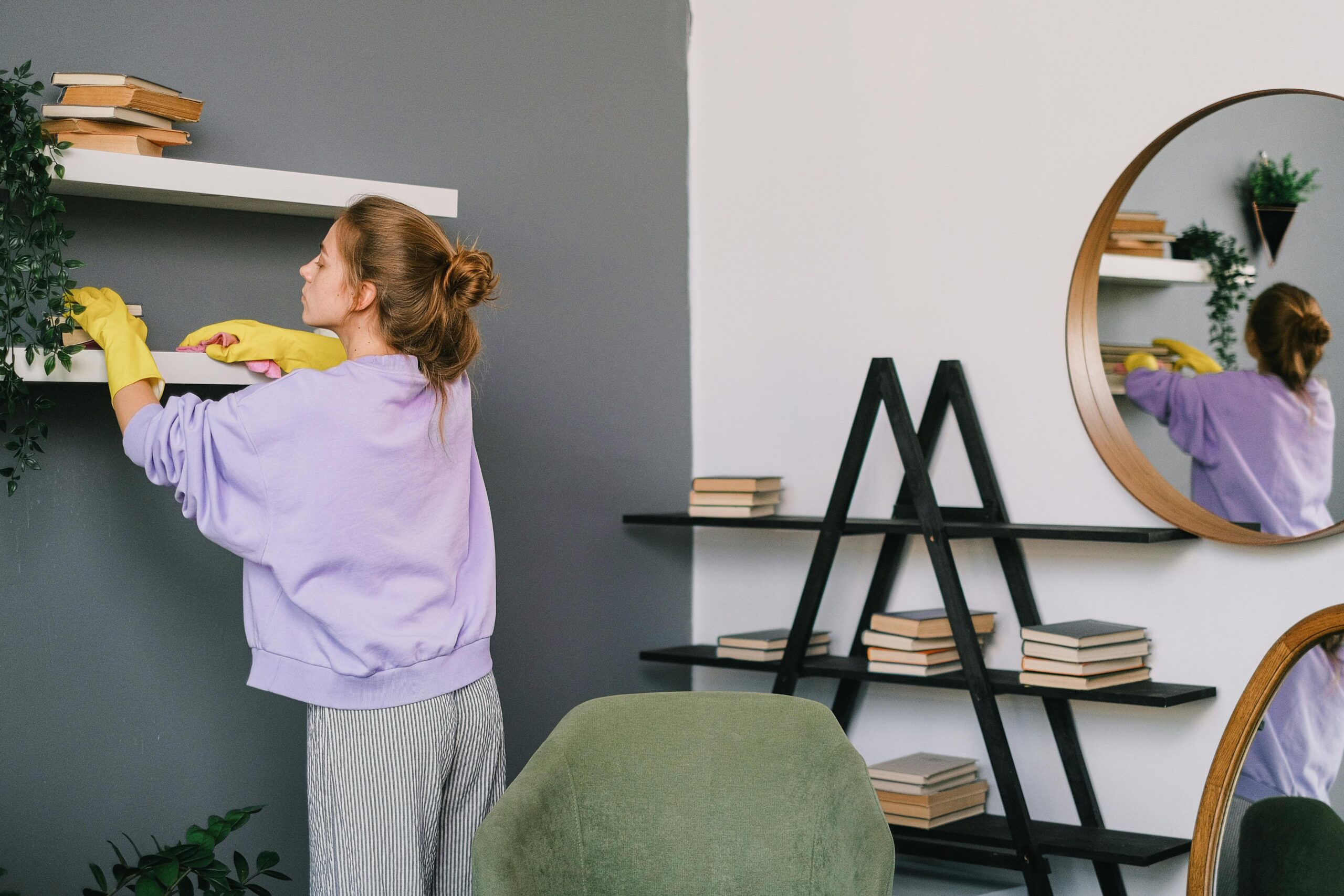A woman cleans shelves in a stylish living room with mirrors and decor.